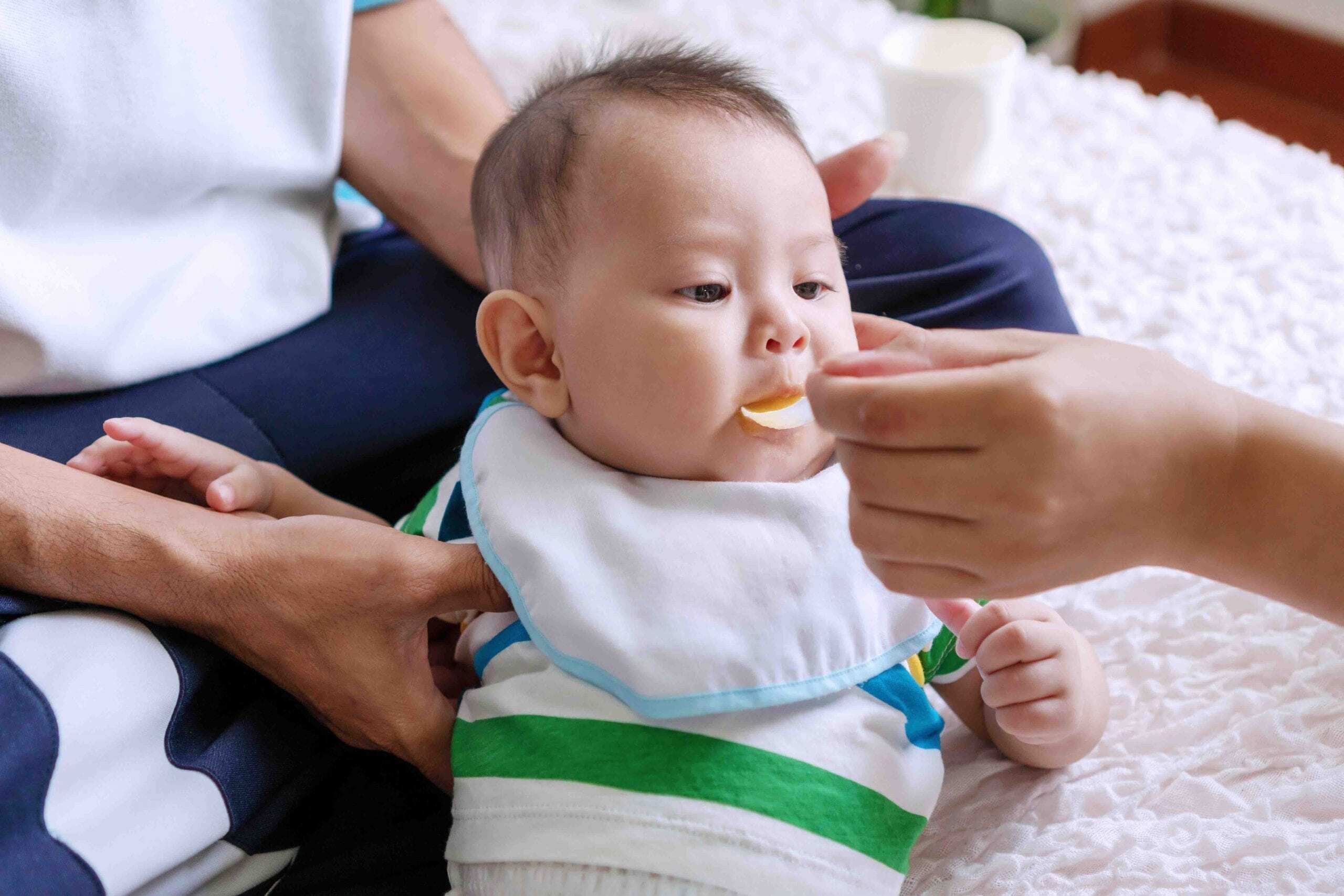 Portrait cute baby boy lying on the lap of his father while hand of his mother holding spoon feeding food for his. Asian young woman feed male for her son. Lifestyle child concept.