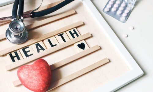 Closeup of red heart stone, stethoscope, wooden letter board and pills on white table background. Health care, medical examination. Heart insurance and cardiology concept. World Health day.