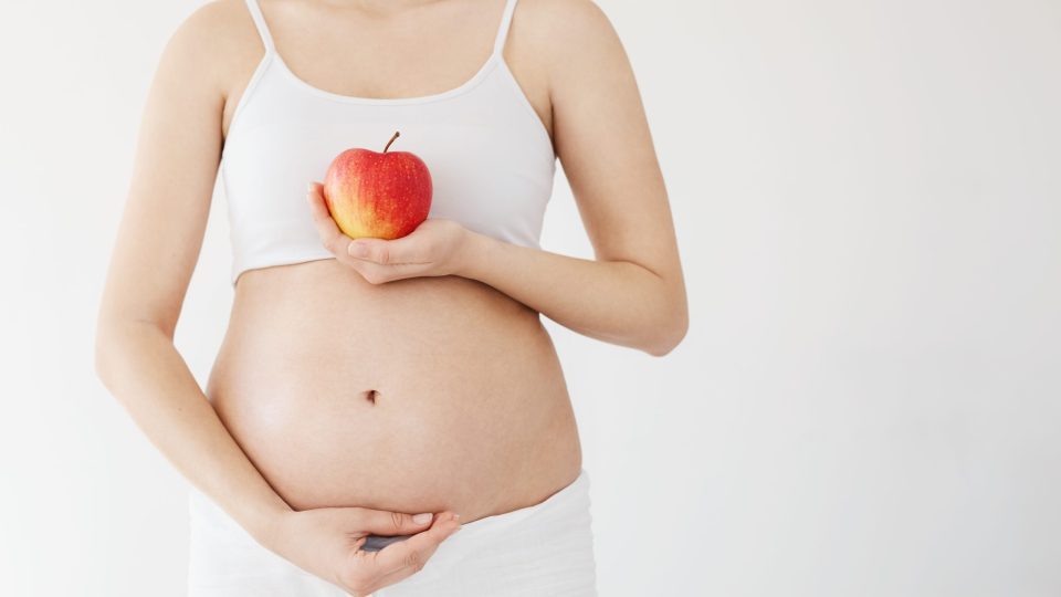 Front portrait of healthy pregnant woman holding her belly and an apple full of vitamins. Pregnancy healthy diet concept over white.